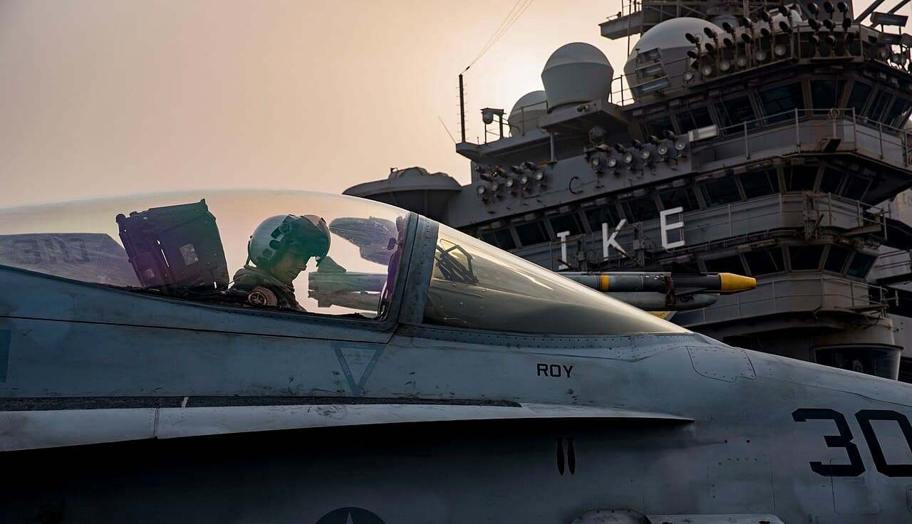 A helmeted pilot sits in the cockpit of an F/A-18C Hornet taxiing across the flight deck of the carrier USS Dwight D. Eisenhower, with the ship's island behind